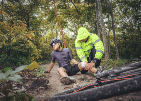 ciclista de bmx sentado no meio do monte, rodeado de árvores, com o joelho magoado e um homem de amarelo a fazer o curativo.